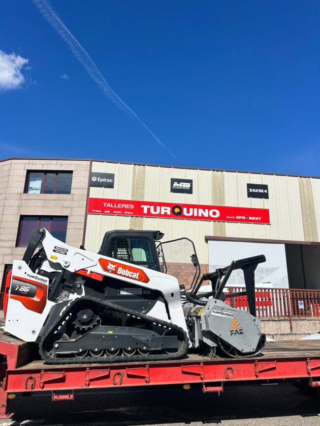 Bobcat T86 con implemento FAE en Talleres Turquino Cargadora de cadenas Bobcat T86 con implemento FAE sobre una plataforma de transporte, frente a las instalaciones de Talleres Turquino.
